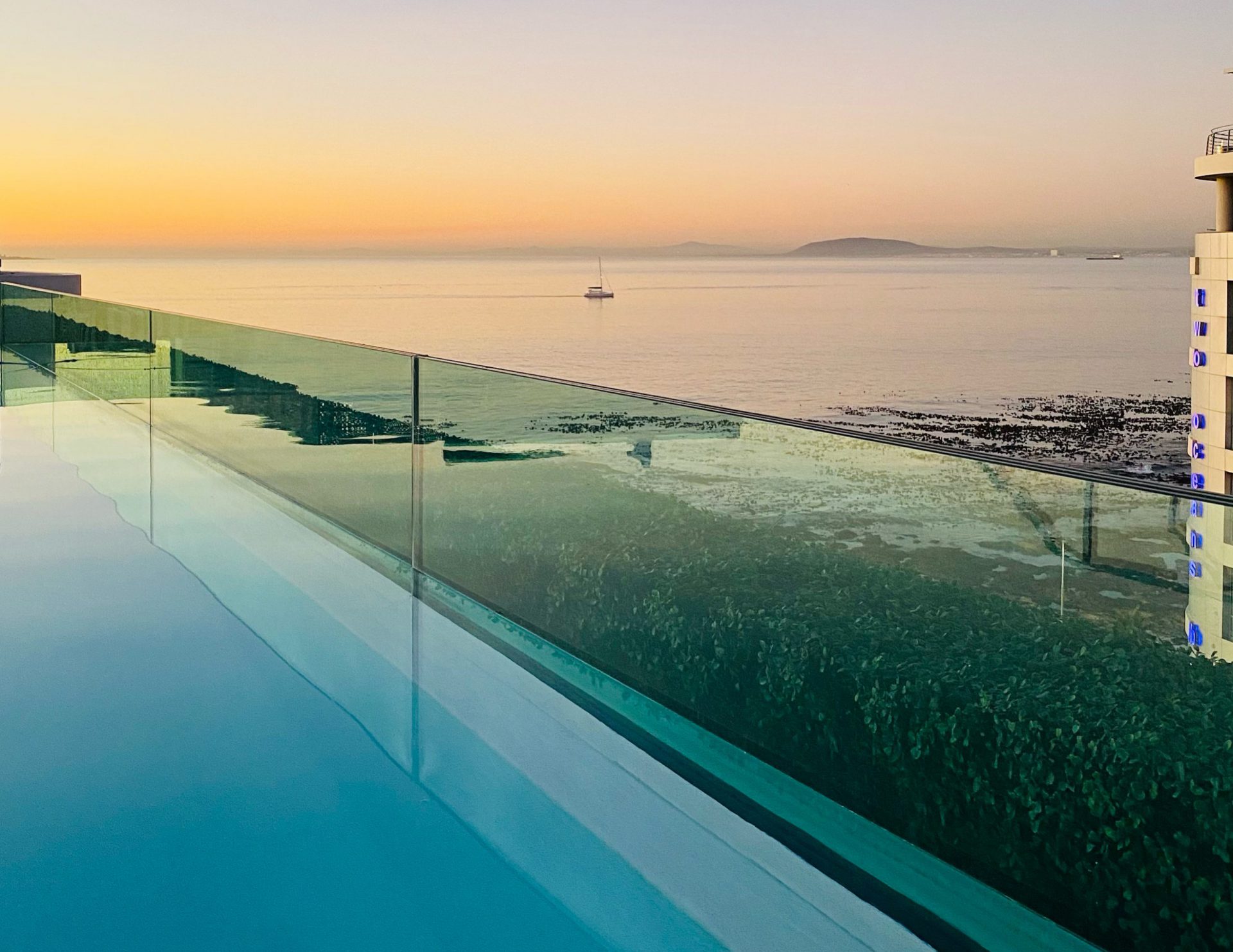 A view of a sunset over the ocean from a long, clear blue pool bordered by a transparent green glass screen.