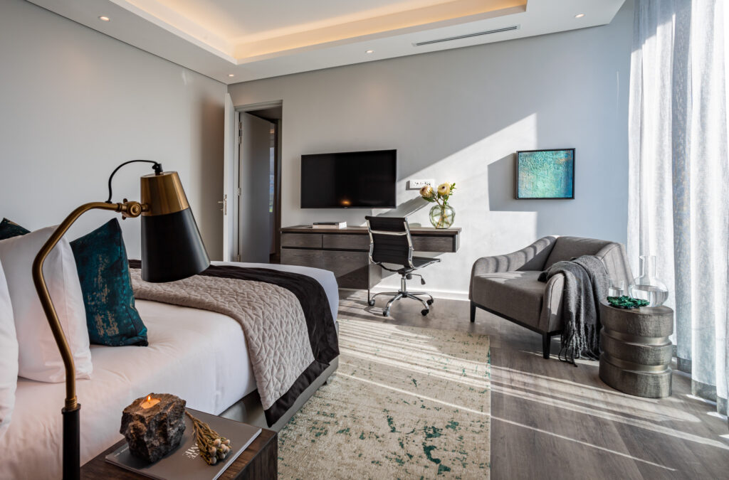 The interior of a bedroom with a double bed looking onto a desk and tv, with seating space and light pouring in from windows on the right hand side