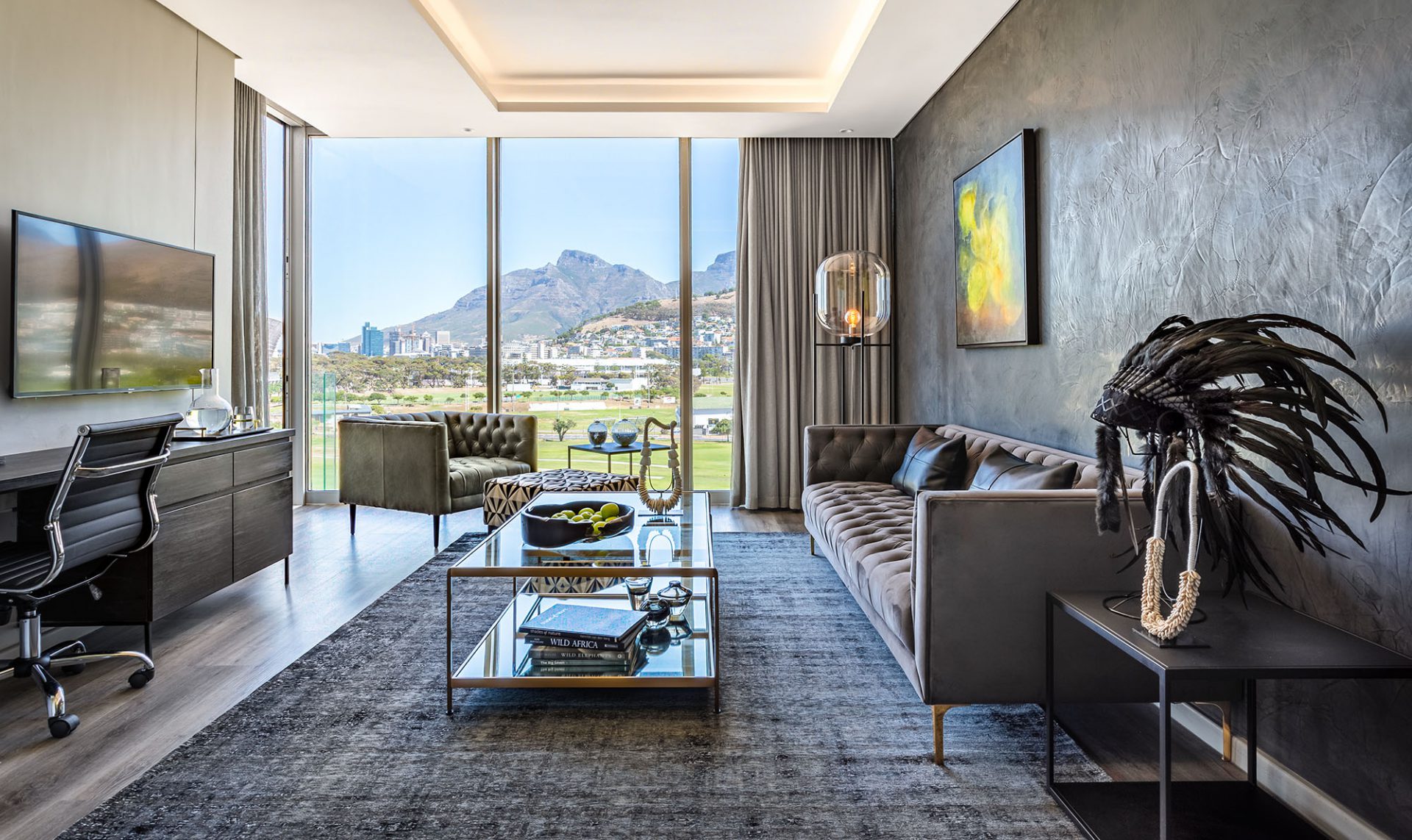 A luxurious living room setting with dark finishes and floor to ceiling windows that look out onto a golf course. The room has a couch, coffee table, lamp, television, workstation and a canvas on the wall.