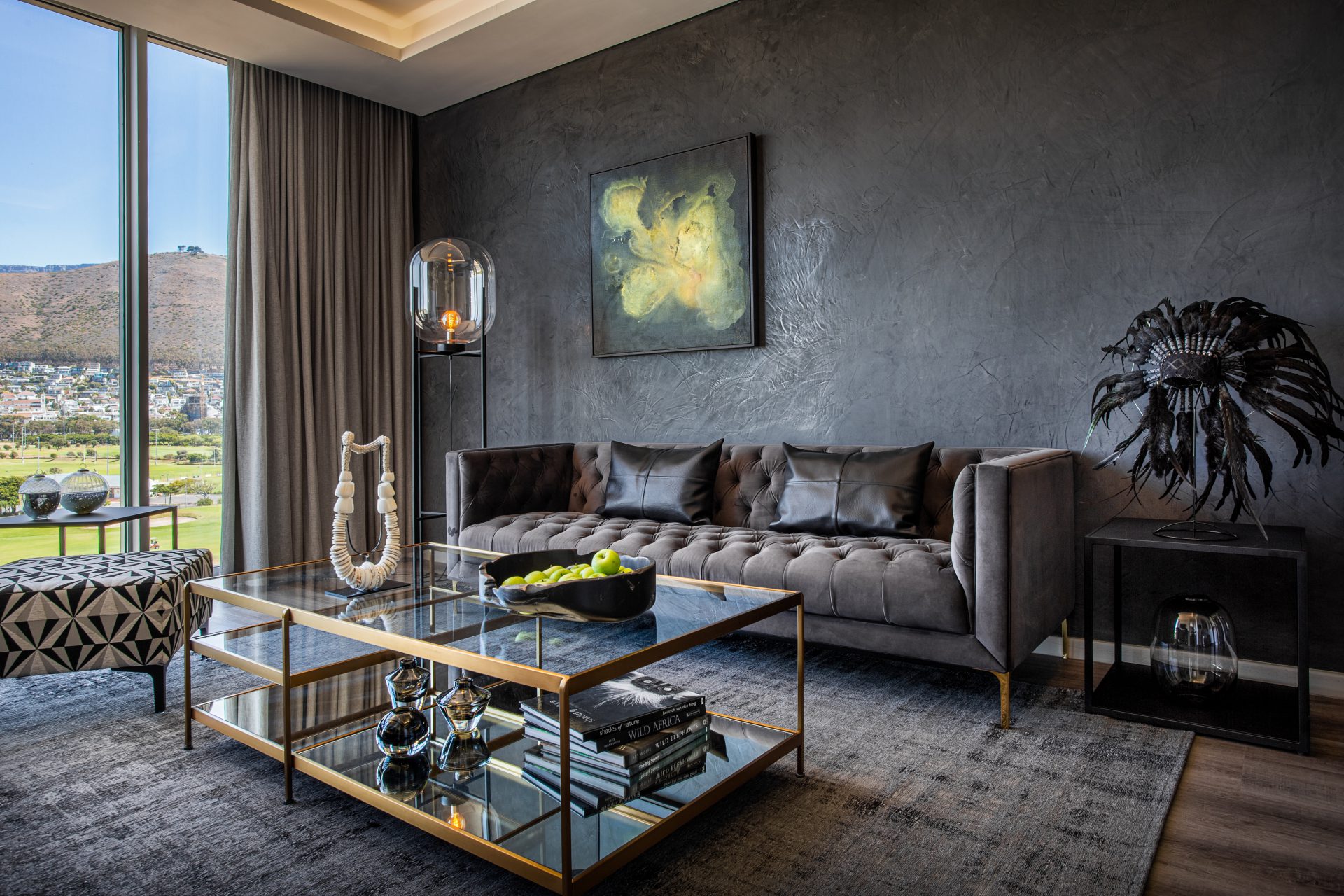 A luxurious living room setting with dark finishes and floor to ceiling windows that look out onto a golf course. The room has a couch, coffee table, lamp and a canvas on the wall.