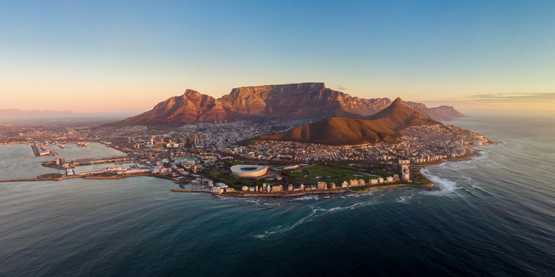 A helicopter view of Cape Town at sunset, displaying the mountains, city centre, Green Point stadium and ocean around it