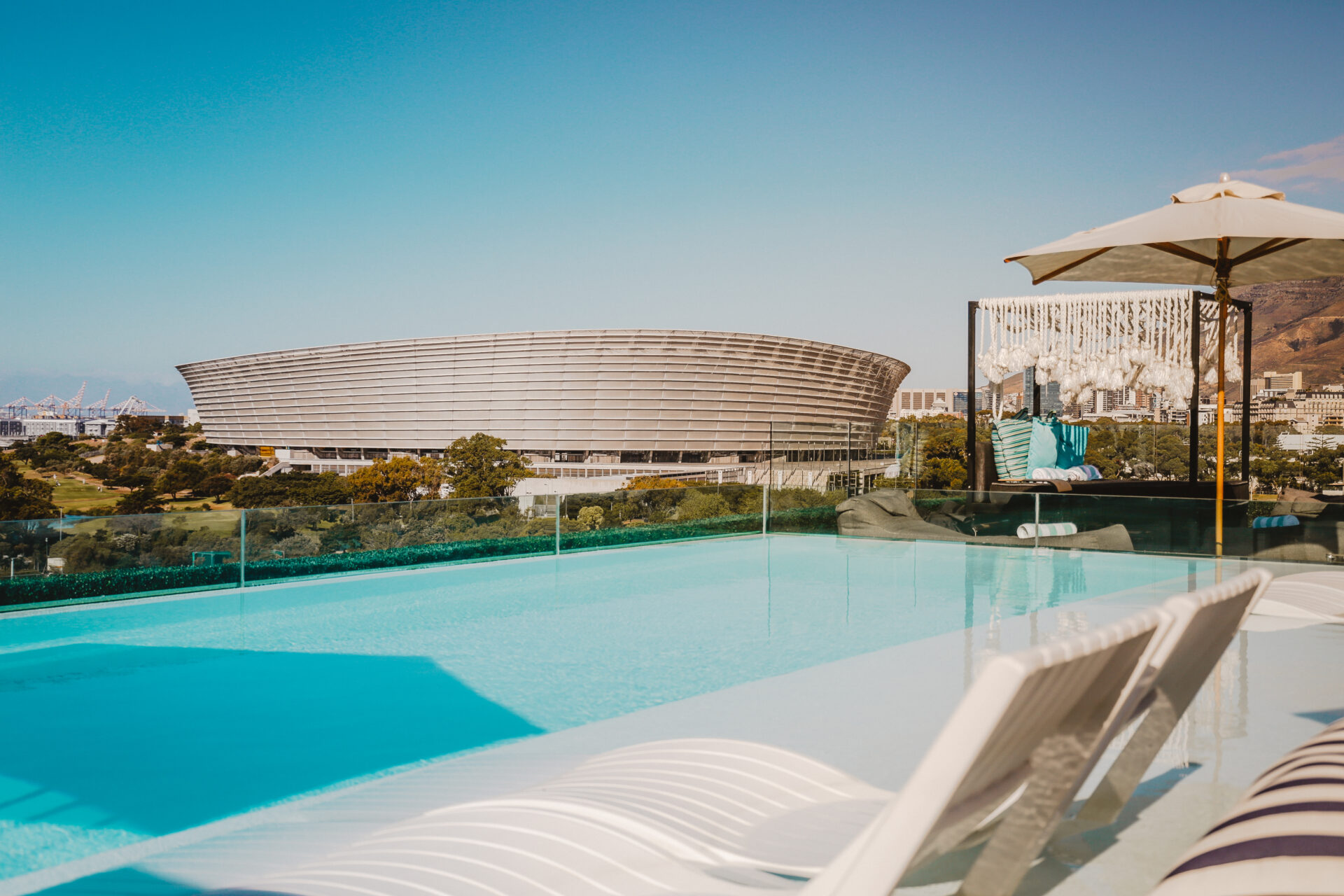 A view of the Greenpoint Stadium on a sunny day from a balcony with a long, clear blue pool, lounging chairs and an umbrella